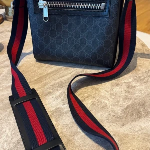 Gucci axelväska med GG-mönster - Snygg svart axelväska från Gucci med klassiskt GG-mönster och bred axelrem i rött och marinblått. Väskan har silverfärgad dragkedja och detaljer, samt en praktisk ytterficka. Tillverkad i canvas med skinninslag och märkning på insidan.
