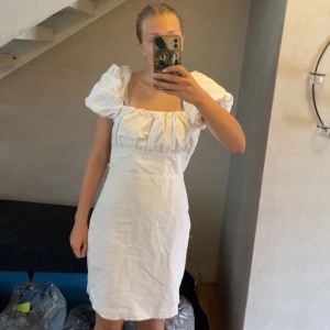 Vit kort klänning med puffärmar - Superfin vit kort klänning med puffiga ärmar och snygg snörning i ryggen. Klänningen har fyrkantig urringning, figurnära passform och dragkedja i sidan. Perfekt för sommarens alla tillfällen och ger en romantisk vibe.