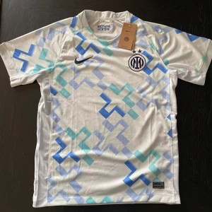 Inter Milan x Nike vit fotbollströja - Inter Milan x Nike fotbollströja i vitt med blå och turkosa geometriska mönster. Broderat Inter-emblem och Nike-logga på bröstet. Tillverkad i lätt och ventilerande polyester, perfekt för match eller träning. Snygg modern design med korta ärmar och rund hals.