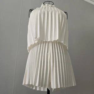 Vit plisserad kortklänning med hög krage - Stilren vit kort jumpsuit i plisserat tyg – ser ut som en klänning men är i själva verket ett par shorts! Ärmlös modell med hög krage och vackert fall. Lätt volang vid halsen och knytning i nacken. Luftig, bekväm och perfekt för sommarens alla tillfällen – både vardag och fest.