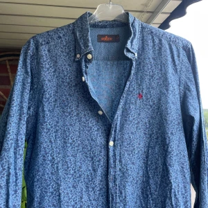 Blommig blå skjorta från Morris - Snygg blå skjorta från Morris med diskret blommigt mönster och klassisk krage. Skjortan har vita knappar och lång ärm, perfekt för dig som vill ha en stilren men ändå unik look. Broderad röd logga på bröstet ger extra detalj.