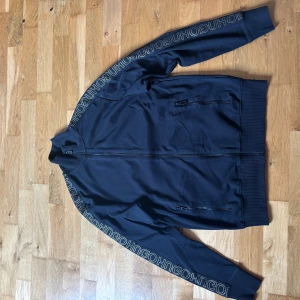 Svart zip-tröja från Hugo Boss - Svart zip-tröja från Hugo Boss med ribbade muddar och dragkedja framtill. Snygg detalj med HUGO-tryck längs ärmarna och två fickor med dragkedja. Storlek XS men passar även S. 