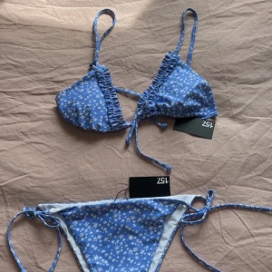Blå blommig bikini från 157 - Supersöt blå bikini med vita små blommor från 157. Klassisk trekantsmodell med knyt både upptill och i sidorna på underdelen. Perfekt för stranden eller poolen, och den blommiga designen ger en fräsch sommarkänsla. Aldrig använd! 