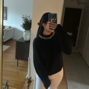 Svart stickad tröja med kedjedetalj - Svart stickad tröja med rund hals och en chunky guldfärgad kedja runt halsringningen. Tröjan har lång ärm och en loose fit som ger en chill vibe. Perfekt för dig som gillar att sticka ut med detaljer och vill ha något både bekvämt och snyggt.