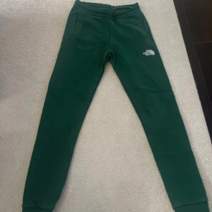 Mörkgröna mjukisbyxor The North Face - barn 13 år  - Mörkgröna mjukisbyxor från The North Face med vit logga på låret. Byxorna har resår i midjan och vid bensluten samt två sidofickor. Perfekta för chill eller träning, tillverkade i mjukt material för extra komfort.