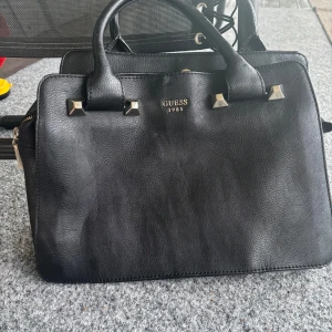 Svart handväska från Guess i skinn - Snygg svart handväska från Guess med stilrena gulddetaljer och logga framtill. Väskan har två handtag, flera fack och dragkedja i mitten. Tillverkad i skinn med en klassisk, boxig form som passar till många outfits. Liten sliten se bilderna❤️