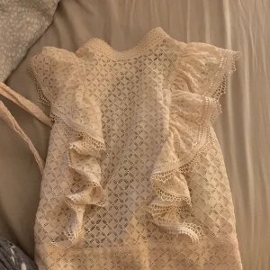 Vit spetsblus med volanger Bik Bok - Supersöt vit blus från Bik Bok i spets med genombrutet mönster och volangdetaljer längs axlar och framstycke. Blusen har korta ärmar, knäppning i nacken och knytband i midjan. Perfekt för dig som gillar romantisk och feminin stil.