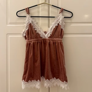 Rosa sammetslinne med spetsdetaljer - Supersött linne i mjuk rosa sammet med vita spetsdetaljer längs v-ringningen och nederkanten. Smala axelband och lätt rynkad midja ger en romantisk vibe. Perfekt för dig som gillar en feminin och trendig look. Storlek S 