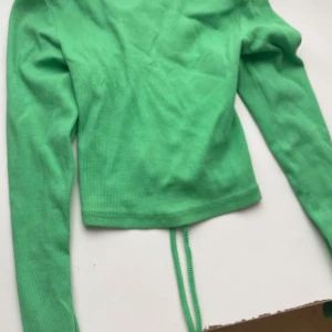 Grön ribbad långärmad topp med snörning - Säljer en grön ribbad långärmad topp med snygg snörning i ryggen. Toppen är croppad och har en tight passform som framhäver figuren. Perfekt för dig som vill sticka ut med en cool detalj på ryggen. Materialet är mjukt och stretchigt för extra komfort.