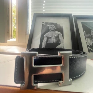  skärp med H-spänne - Snyggt svart med ikoniskt H-spänne i silverfärgad metall. Bältet är gjort i äkta skinn med vit kontrastsöm längs kanterna. Perfekt accessoar för att lyfta din outfit med en lyxig touch.