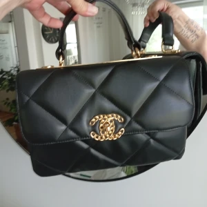 Svart quiltad handväska - Säljer en svart handväska med klassiskt quiltat mönster och guldfärgade detaljer. Väskan har ett snyggt handtag och den ikoniska loggan i guld framtill. Perfekt för dig som vill ha en lyxig och tidlös accessoar.