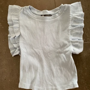 Ljusblå ribbad topp med volangärmar Zara - Supersöt ljusblå ribbad topp från Zara med volangdetaljer på axlarna. Toppen är ärmlös och har en rund halsringning. Perfekt för dig som gillar en  trendig stil med extra detaljer på axlarna.