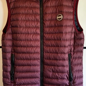 Vinröd dunväst från Colmar - Snygg vinröd dunväst från Colmar med quiltad design och svarta dragkedjor. Västen har rund halsringning, två fickor med dragkedja och ett diskret Colmar-märke på bröstet. Perfekt att slänga över en hoodie eller tröja när det är kyligt ute. Finns en liten fläck på baksidan som man kan se på bilden men inget märkbart direkt!