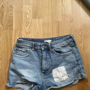 Blå jeansshorts med patch - Snygga blå jeansshorts med råa kanter och en vit patch på ena benet. Klassisk femficksmodell med bälteshällor och slitna detaljer för en cool, avslappnad stil