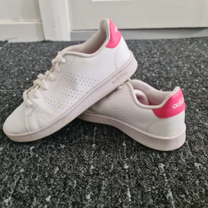 Vita Adidas sneakers med rosa detalj - Snygga vita Adidas sneakers med klassisk design och perforerade sidor. Skorna har en rosa detalj på hälen med Adidas-logga och är tillverkade i syntetmaterial. Perfekta för en clean och sportig look.