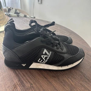 Svarta sneakers från Emporio Armani EA7 - Svarta sneakers från Emporio Armani EA7 med meshpaneler och detaljer i syntet. Snygg logga på sidan och bak, samt svarta snören. Skorna har en sportig vibe med platt sula och rund tå. Perfekta för dig som gillar stilrena och bekväma sneakers.