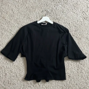 Svart t-shirt med knyt från Nelly - Svart t-shirt från Nelly i storlek XS. Modellen har korta ärmar, rund halsringning och en snygg knyt-detalj i ryggen som ger en cool twist. T-shirten är i mjuk bomull och har en lite loose passform. Perfekt för dig som gillar stilrena basplagg med en extra detalj.❣️❣️