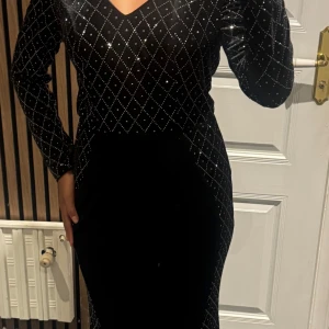 Svart långklänning med glittriga detaljer - Säljer en svart långklänning med v-ringning och långa ärmar. Klänningen är täckt av ett glittrigt rutmönster med små pärlor som ger en lyxig känsla. Materialet är mjuk sammet som sitter snyggt och har en elegant siluett. Perfekt för dig som vill sticka ut på festen!!! Man får ha i åtanke att jag är kort (160) ) så kolla så att den passar först !