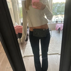 Svart glittrig axelväska med kedja i jeansmaterial - Snygg liten svart axelväska med glittriga detaljer och silverfärgad kedja som axelrem. Väskan har en rektangulär form och dragkedja upptill. Perfekt för att lyfta din outfit med lite extra bling. Den är i svart grått jeansmaterial!!