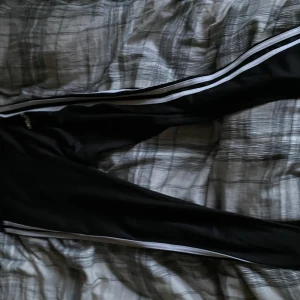 Svarta Adidas mjukisbyxor med vita ränder - Svarta mjukisbyxor från Adidas i storlek M med klassiska vita ränder längs sidorna och Adidas-logga på höften. Byxorna är gjorda i polyester och elastan, har en slim passform och är perfekta för träning eller chill. Snygg sportig look med bekväm känsla.