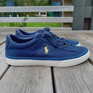 Ralph Lauren skor - Snygga marinblå sneakers från Polo Ralph Lauren med klassisk gul logga på sidan. Skorna har ovandel i canvas, vita sulor och matchande blå snörning. Perfekta för en clean och stilren look, med Polo-detaljer på både plös och häl.