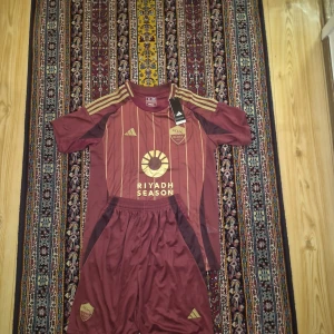 AS Roma hemmaställ 2024/25 - Snygg AS Roma fotbollset från Adidas i vinröd med guldiga ränder och detaljer. Kortärmad, slim fit och tillverkad i lätt polyester. Tröjan har klubbmärke, Adidas-logga och 'Riyadh Season' tryck på bröstet. Perfekt för dig som vill sticka ut på planen.