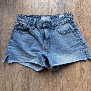 Blå jeansshorts från Cubus - Snygga blå jeansshorts från Cubus i klassisk femficksmodell. De har hög midja, råa kanter och slitna detaljer för en cool vibe. Materialet är robust denim med ljus tvätt och knappstängning framtill. Perfekta för varma dagar och chill stil.