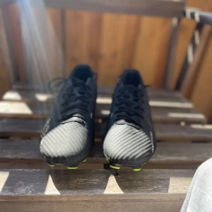 Nike Air Zoom fotbollsskor svart/gul - Säljer ett par Nike Air Zoom fotbollsskor i svart med detaljer i silver och neon-gult. Skorna har snörning, låg profil och en platt sula med dobbar för gräsplan. Snyggt mönstrad ovandel i syntetmaterial och Nike-logga på sidan. Perfekt för dig som vill ha grymt grepp och stil på planen.