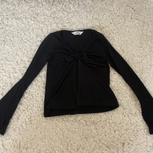 Svart långärmad topp med rosett H&M - Svart långärmad topp från H&M med snygg rosettdetalj framtill. Modellen har en djupare ringning och är tillverkad i mjukt material som känns skönt mot huden. Perfekt för dig som gillar stilrena och trendiga plagg med en twist.