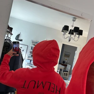 Röd Helmut Lang hoodie  - Säljer en röd hoodie från Helmut Lang med svart texttryck både fram och bak. Tröjan har huva med metallöljetter och en stor magficka. Väldigt sällsynt och perfekt för dig som gillar streetwear och vill sticka ut. Skriv gärna och fråga om bilder och diskutera pris!🤝🏻🤝🏻✨