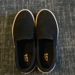 Svarta slip-on sneakers från 157 - Svarta slip-on sneakers från 157 med vit sula och klassisk rund tå. Skorna är i textilmaterial och har en enkel, stilren design utan snörning. Perfekta för en avslappnad och clean look.