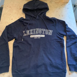 Mörkblå hoodie från Lexington - Snygg mörkblå hoodie från Lexington med stor vit logga på bröstet och snörning i huvan. Skön bomull på insidan. Perfekt för en chill dag. Bra passform. Använd fåtal gånger