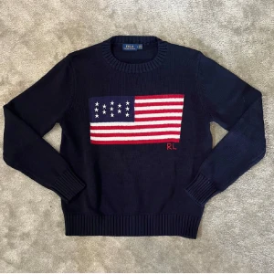 Mörkblå stickad tröja Polo Ralph Lauren - Snygg mörkblå stickad tröja från Polo Ralph Lauren med amerikansk flagga i rött, vitt och blått på bröstet samt RL-broderi. Rund hals och ribbade muddar vid ärmslut och nederkant. Perfekt för dig som gillar klassisk och cool stil. Fick den som present men den är tyvärr förliten, har inget kvitto så gick inte att lämna tillbaks så säljer för en snabb peng