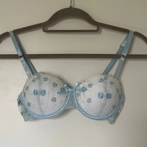 Blommig ljusblå blus med spetsdetaljer - Superfin vit BH med ljusblå broderade blommor i blått och vitt samt spetsdetaljer längs kanterna i storlek 70B. Tunna justerbara axelband och en liten rosett framtill. Med följer även ett par matchande stringtrosor. Aldrig använda🩵