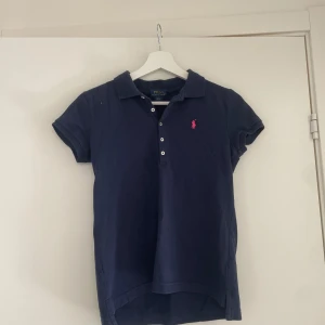 Mörkblå Polo Ralph Lauren t-shirt - Snygg mörkblå t-shirt från Polo Ralph Lauren med klassisk pikékrage och korta ärmar. T-shirten har knappar framtill och en rosa broderad logga på bröstet. Tillverkad i mjuk bomull som är skön mot huden. Perfekt för dig som gillar stilren och sportig look.