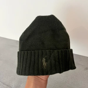 Mörkgrön stickad mössa Polo Ralph Lauren - Snygg mörkgrön stickad mössa från Polo Ralph Lauren med klassisk broderad logga framtill. Mössan har bred uppvikt kant i ribbstickat material och är perfekt för kyliga dagar. Enkel och stilren design som funkar till allt.