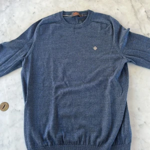 Blå merinoulltröja från Morris - Snygg blå tröja från Morris i 100% merinoull. Klassisk rund hals och diskret broderad logga på bröstet. Tröjan har långärmade och ribbade muddar vid ärmslut och nederkant. Perfekt för dig som gillar stilrena och bekväma plagg.