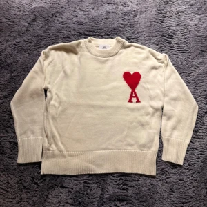 Vit stickad tröja Ami Paris hjärta - Vit stickad tröja från Ami Paris med röd hjärtlogga och bokstaven A på bröstet. Klassisk rund halsringning, ribbade muddar och nederkant. Perfekt för dig som gillar stilrena plagg med en twist. Köpt på hemsidan