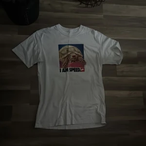 Vit Nike t-shirt med sköldpaddstryck - Vit t-shirt från Nike med ett stort tryck av en sköldpadda och texten 'I AM SPEED' på bröstet. Klassisk passform med rund hals och korta ärmar. Perfekt för dig som gillar humoristiska motiv och streetwear.