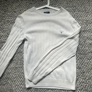 Vit ribbad tröja från GANT - Snygg vit tröja från GANT med diskret logga broderad på bröstet. Tröjan har ett ribbat mönster och är långärmad med rund hals. Materialet är mjuk bomull som känns skönt mot huden. Perfekt för dig som gillar klassisk och stilren design. Hål i ärmen, se bild 2
