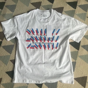 Maison Margiela vit t-shirt med tryck - Maison Margiela t-shirt i vit bomull med coolt grafiskt tryck i blått och rött framtill. Klassisk rund hals och korta ärmar. Perfekt för dig som gillar statement-plagg och designer vibes.
