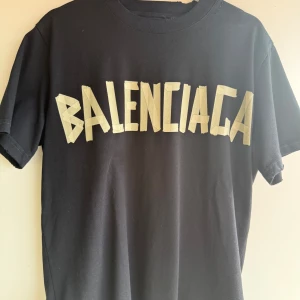 Svart Balenciaga t-shirt oversized - Cool svart t-shirt från Balenciaga med stort beige tryck på framsidan och gul text samt BB-logga på ryggen. T-shirten har klassisk rund hals och korta ärmar. Materialet känns som mjuk bomull och passformen är oversized. Perfekt för dig som gillar statement-plagg och streetstyle.