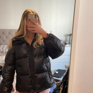 Svart pufferjacka - Säljer en svart pufferjacka med huva och dragkedja. Jackan är riktigt fluffig och har en glansig finish. Perfekt för kalla dagar och har en snygg, oversized look. Passar dig som vill ha en stilren och trendig vinterjacka.