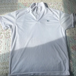 Vit pikétröja från Lacoste - Vit pikétröja från Lacoste med klassisk krage och knappar framtill. Ikoniska gröna krokodilloggan på bröstet. T-shirten är kortärmad och har en stilren, sportig look. Perfekt för dig som gillar enkel och tidlös stil.