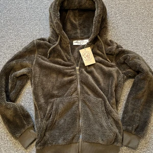 Mörkgrön fleecehoodie Amaci & Sons - Supermjuk och mysig mörkgrön hoodie från Amaci & Sons i fleece. Tröjan har huva med snörning, dragkedja framtill och ribbade muddar vid ärmslut och nederkant. Perfekt för kyliga dagar när du vill vara både bekväm och snygg.