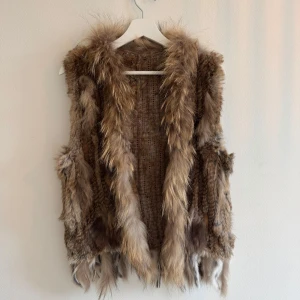 Brun pälsväst i äkta päls - Säljer en fluffig och cool brun pälsväst i äkta päls. Västen har en naturlig mix av ljusbruna och mörkbruna toner, långhårig päls och snygga detaljer längs kanterna och nederdelen. Kan knäppas med 1 knapp fram