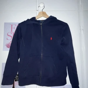Mörkblå hoodie från Polo Ralph Lauren - Snygg mörkblå hoodie från Polo Ralph Lauren med röd broderad logga på bröstet. Tröjan har huva, dragkedja framtill och två fickor. Tillverkad i mjuk bomull, perfekt för chill dagar. Klassisk och stilren design som alltid funkar. Storleken vet ja ej hur ja ska tolka men skulle säga xxs. Nypris runt 1100