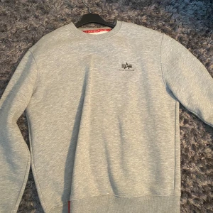 Grå sweatshirt från Alpha Industries - Snygg grå sweatshirt från Alpha Industries med diskret logga på bröstet. Tröjan har rund hals, långa ärmar och ribbade muddar vid ärmslut och nederkant. Perfekt för chill dagar eller när du vill ha en clean look.