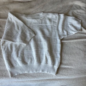Vit stickad kortärmad tröja - Vit stickad tröja med korta ärmar och ribbade muddar. Tröjan har ett diskret strukturmönster i ränder och en rund halsringning. Mycket fint skick och 