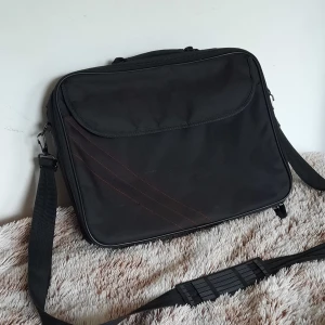 Svart datorväska med axelrem - En svart datorväska i slitstark polyester med justerbar axelrem och praktiskt innerfack. Väskan har lock med röda dekorativa sömmar och dragkedja runt om. Perfekt för att bära laptop och tillbehör till plugget eller jobbet. Ett hål finns (Bild 4) men de påverkar inte väskan ett dugg. Pris kan diskuteras  Skriv om ni har funderingar! :)